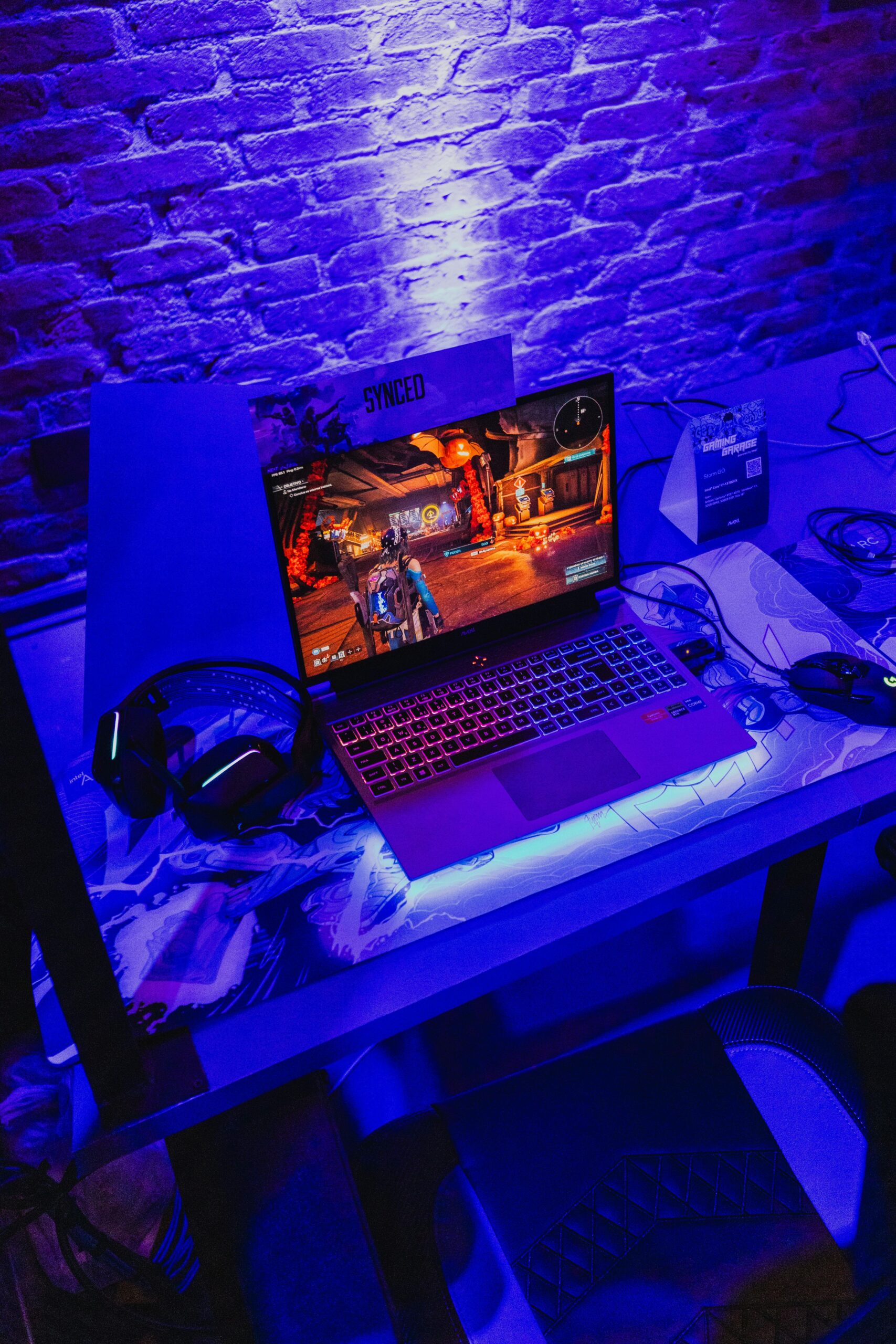 Modern gaming laptop with headphones and mouse on desk in purple neon lighting.