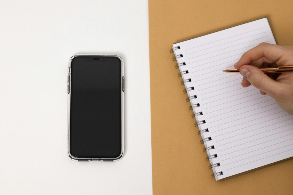 Minimalist top view of a workspace setup featuring a smartphone and notebook with a hand holding a pen.