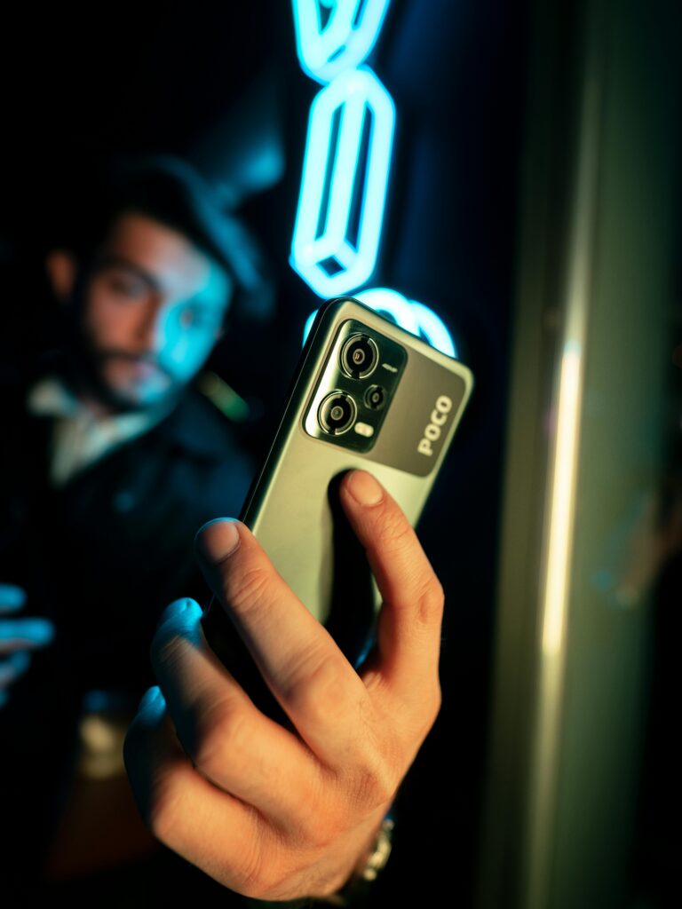 A man holds a Poco smartphone in a dimly lit indoor setting, with neon lights in the background.