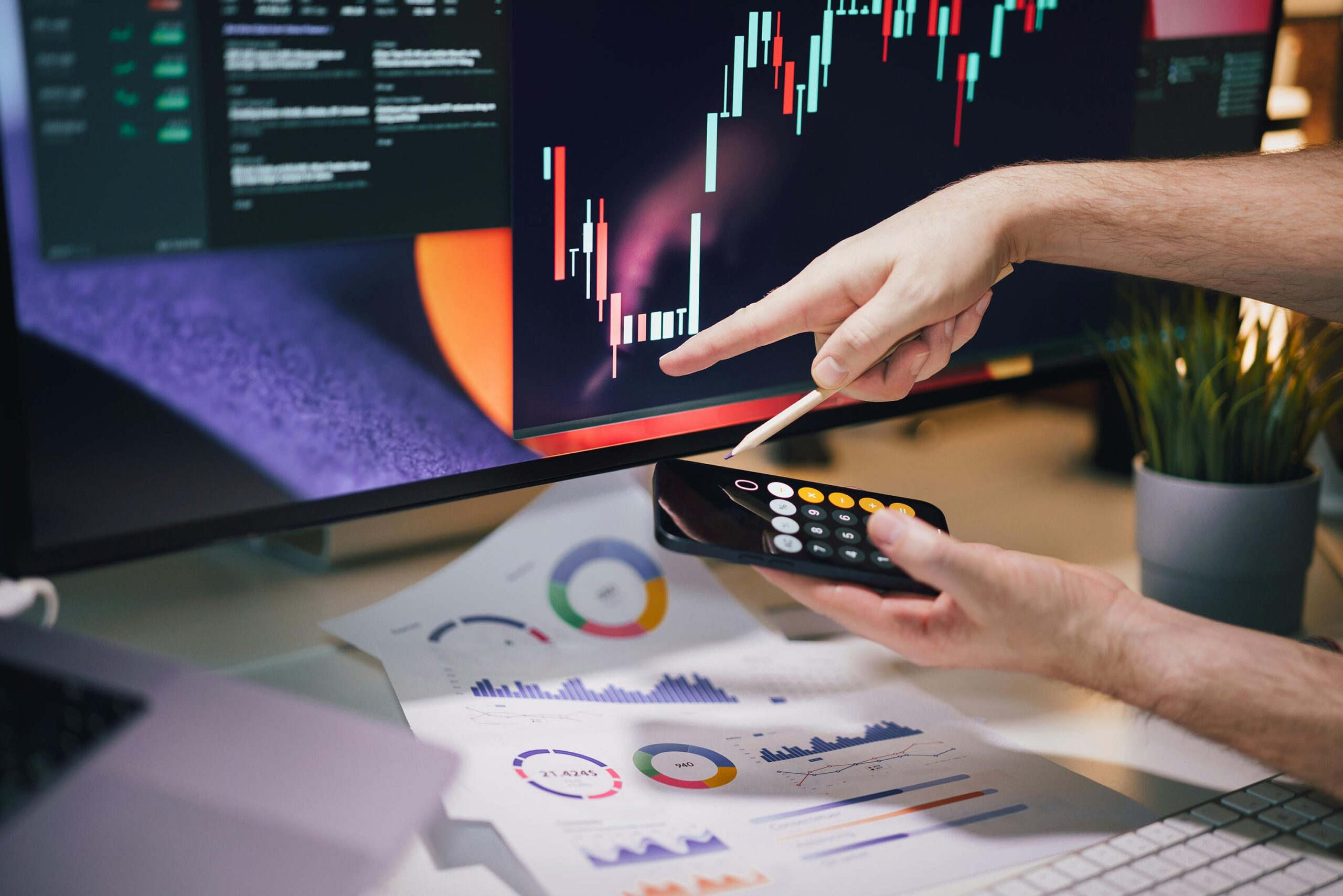 Hands analyzing stock market data on monitor and smartphone in office setting.